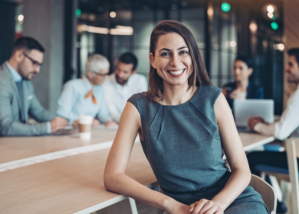 A strong female leader smiling and facing the camera with her team at a desk in the office blurred in the background.
